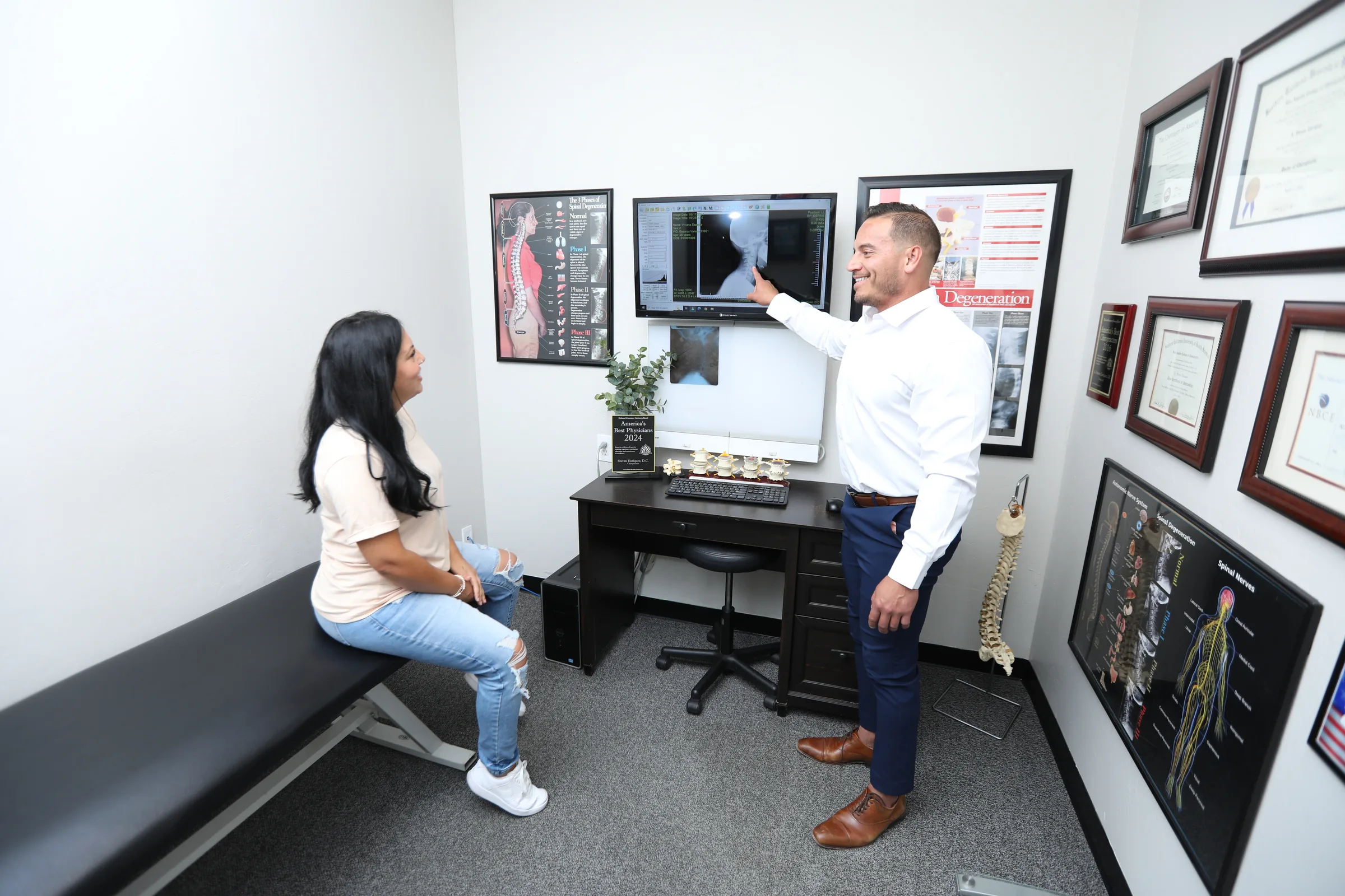 Chiropractor in white shirt pointing to X-ray on monitor while patient sits on examination table.